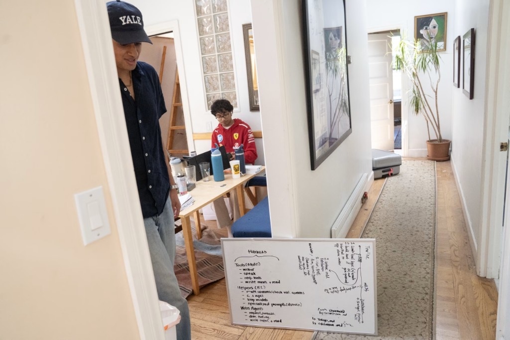 Ariyan in the hallway doorway beside a whiteboard labeled Harness, with a resident in a red hoodie working at the table
